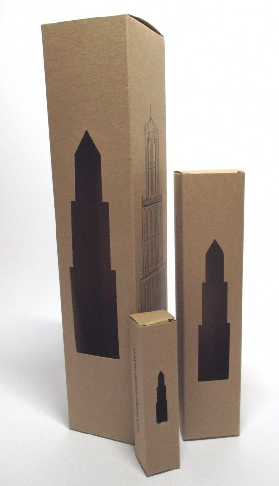 Drie verschillende formaten bruine kraft kartonnen doosjes voor kaarsen, bedrukt in zwart, elk voorzien van een venster in de vorm van een kerktoren silhouet de Dom toren van Utrecht. Deze verticale kaarsenverpakkingen tonen de veelzijdigheid van Bedrukte-doosjes.nl in het maken van thema-verpakkingen voor souvenirkaarsen en lange tafelkaarsen met een unieke stansvorm met speciaal venster.