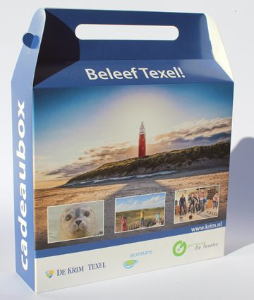 Een rechthoekige, op maat gemaakte kartonnen doos met handvat, uitgevoerd als luxe welkomstpakket voor een camping op Texel. De verpakking is volledig bedrukt in full color met een sfeervolle landschapsfoto van het Texelse strand bij zonsondergang, inclusief de iconische rode vuurtoren. Boven de foto staat in witte letters de uitnodigende tekst 'Beleef Texel!'. De onderkant van de doos toont drie kleinere detailfoto's: een close-up van een zeehond, spelende kinderen in de duinen en een sfeerbeeld van gasten, geflankeerd door logo's van partners zoals De Krim Texel en Ecomare. De zijkant is voorzien van de tekst 'cadeaubox' in verticale witte letters tegen een blauwe achtergrond. Het geïntegreerde handvat aan de bovenzijde maakt dit vouwkartonnen doosje tot een handige en gewaardeerde goodie bag om uit te delen aan gasten bij aankomst op het event of de vakantiebestemming.