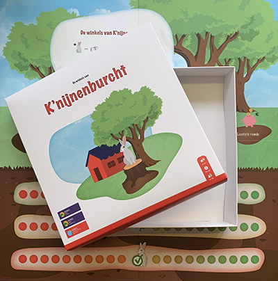 Een op maat gemaakte, wit bedrukte speldoos voor het spel 'K'nijnenburcht', liggend op een groot, kleurrijk bijbehorend spelbord met een natuurthema. De afbeelding toont de geopende doos met een illustratie van een konijn bij een burcht, geproduceerd door Drukkerij Van der Louw. Dit is een voorbeeld van een complete spelset waarbij zowel de spelverpakking als het spelbord zijn bedrukt in eigen drukkerij voor een professionele uitstraling. De doos is een vierkante speldoos met los deksel