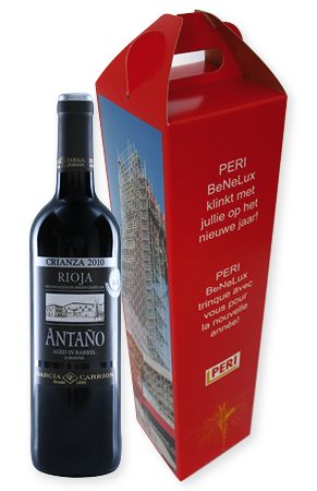 Een op maat gemaakte rode kartonnen doos met handvat, specifiek ontworpen voor een fles wijn. Naast de verpakking staat een volle fles Antaño Rioja Crianza wijn. De doos is bedrukt met een grote, gedetailleerde afbeelding van een steigerconstructie op de zijkant. De voorzijde bevat een tweetalige pakkende tekst in het wit: 'PERI BeNeLux klinkt met jullie op het nieuwe jaar!' en de Franse vertaling 'PERI BeNeLux trinque avec vous pour la nouvelle année!', inclusief het PERI-bedrijfslogo. De verpakking is voorzien van een stevig, geïntegreerd handvat aan de bovenzijde en toont onderaan een subtiele afbeelding van vuurwerk. Dit model illustreert hoe een doos met handvat van extra stevig vouwkarton wordt gebruikt als zakelijke geschenkverpakking voor zwaardere producten zoals flessen, waarbij full color bedrukking zorgt voor een premium uitstraling.