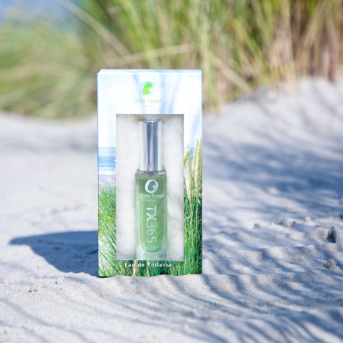 Een medium close-up van een bedrukt schuifdoosje voor het O'de Texel parfum, gefotografeerd op een zandduin, wat de natuurlijk geïnspireerde uitstraling van de verpakking benadrukt. Setting en Sfeer: De foto is genomen bij daglicht, met het doosje geplaatst op licht, fijn zand. Op de achtergrond is wazig duingras zichtbaar, wat de geur geïnspireerd op de Texelse natuur visueel ondersteunt. Parfumdoosje: Het is een langwerpig, smal, kartonnen schuifdoosje. Zowel de sleeve als het doosje zijn in full color bedrukt. De opdruk toont een rustig strand- en duinlandschap met een blauwe hemel en groen gras. Venster met Folie: De sleeve van het doosje heeft een vierkant venster met folie, waardoor het parfumflesje zichtbaar is. Product: Achter het venster is een slank, glazen parfumflesje met een zilveren verstuiver zichtbaar. De geelgroene vloeistof in het flesje is duidelijk te zien, en het etiket draagt de merknaam 