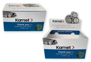 Productfoto van twee op maat gemaakte, bedrukte displaydozen voor het merk Kamet, die fungeren als voorbeeld van professionele toonbank displays en balie displays. De afbeelding toont twee stadia van de verpakking: links een gesloten doos klaar voor transport, en rechts dezelfde doos die is getransformeerd tot een geopende display box met een opvallend pop-up element. De displaydozen zijn uitgevoerd in een strak wit ontwerp met een brede donkerblauwe baan aan de onderzijde. Op de voorzijde staat prominent het zwarte logo 'Kamet >' met de tekst 'Thank you for doing business with us' en een afbeelding van een groene kabelspoel. De geopende variant aan de rechterzijde toont een die-cut pop-up achterkaart (topkaart) die boven de doos uitsteekt. Op deze kaart staan gedetailleerde dwarsdoorsneden van industriële kabels afgebeeld, samen met de slogan 'Your connection to the high temperature world' en de website URL. Deze visuele weergave illustreert een SRP-verpakking (Shelf Ready Packaging) die zowel bescherming biedt tijdens logistiek als directe visuele aantrekkingskracht op de winkelbalie of in het schap, zoals beschreven in de begeleidende tekst over merkpresentatie en verkoopbevordering.