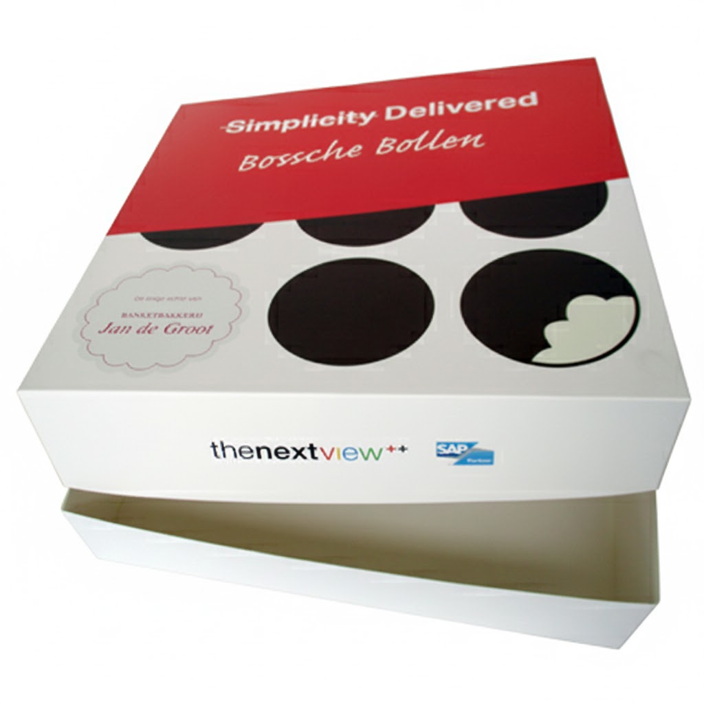 Een luxe, vierkante bedrukte gebaksdoos met een los deksel, specifiek ontworpen voor Bossche Bollen als relatiegeschenk. Het deksel heeft een stijlvol en modern ontwerp: de bovenste helft is helderrood met de tekst 'Simplicity Delivered' en 'Bossche Bollen' in een elegant wit cursief lettertype. De onderste helft van het deksel is wit en gedecoreerd met grote zwarte cirkels die Bossche Bollen symboliseren, waarbij in één cirkel een hapje is 'uitgenomen' om de witte vulling te tonen. Op de linkerzijde van het deksel staat het kwaliteitszegel van 'Banketbakkerij Jan de Groot'. De zijkant van de witte doos is bedrukt met de logo’s van 'thenextview++' en 'SAP Partner', wat de functie van de doos als gepersonaliseerd marketinginstrument voor bedrijven benadrukt. De doos staat op een witte ondergrond met het deksel iets opgetild, waardoor de witte, voedselveilige binnenkant zichtbaar is.