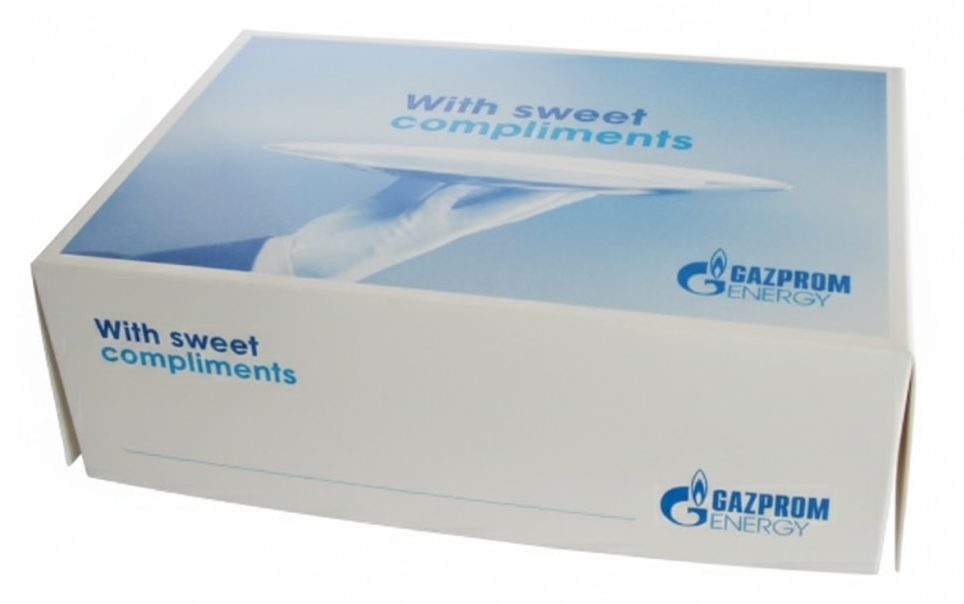 Een close-up in een hoekperspectief toont een vierkante, witte bedrukte gebaksdoos met een los deksel, staand op een witte ondergrond. Dit is een voorbeeld van een bedrukte doos met logo die kan dienen als relatiegeschenk of voor de verpakking van lekkernijen zoals Bossche Bollen, zoals gesuggereerd door de bestandsnaam en de context. De doos is bedrukt in lichte en donkere blauwtinten en wit, wat de stijlvolle en professionele uitstraling benadrukt. Het deksel heeft een full-color opdruk met een subtiel, gradient blauwe hemel- of wolk-achtige achtergrond die overgaat in wit. De belangrijkste visuele elementen op het deksel zijn: De Engelse tekst 'With sweet compliments' in lichtblauwe, cursieve letters, wat de functie als relatiegeschenk en blijk van waardering communiceert. Een afbeelding van een hand, gekleed in een witte handschoen, die een zilveren schaal of dienblad omhoog houdt, wat een gevoel van luxe, service en presentatie oproept—cruciaal voor de merkbeleving van het ingesloten gebak. Het GAZPROM ENERGY-logo met de vlam en de bedrijfsnaam in blauw, prominent rechtsonder op het deksel en herhaald op de voorzijde van de doos. Dit toont hoe een bedrijfslogo duidelijk de afzender van het geschenk communiceert en de herkenbaarheid en meerwaarde vergroot. De bedrukking van deze doos toont een hoogwaardige afwerking, wat past bij de beschrijving van hoogwaardige gebaksdozen bedrukt in full-color die dienen als het visitekaartje van het bedrijf.