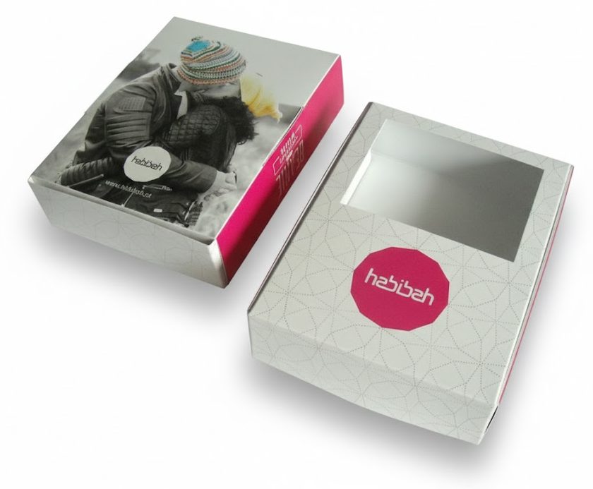 Twee voorbeelden van een bedrukte doos met venster voor het merk Habibah, speciaal ontworpen als verpakking voor modegevoelige artikelen zoals mutsen van biologische wol. De witte vensterdoos op de voorgrond toont een groot rechthoekig uitgesneden venster waardoor de klant de kleur, vorm en maat van de beanie direct kan beoordelen. De zijkant van het doosje is uitgevoerd in een opvallende magenta kleur met de tekst 'Beanie Organic Wool', terwijl de bovenzijde is voorzien van een subtiel geometrisch patroon en het Habibah-logo. De tweede verpakking toont een full-color bedrukking met een lifestyle-afbeelding, wat de juiste uitstraling geeft aan het product. Deze maatwerk productverpakking laat het product aan de klant zien voordat hij het doosje openmaakt, wat de kans op retouren en beschadigde verpakkingen aanzienlijk vermindert.