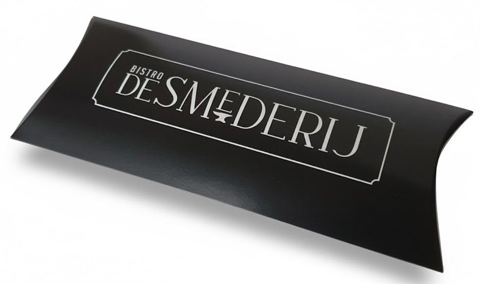 Een elegant, mat zwart bedrukt gondeldoosje (pillow box) met de afmetingen 220x110x30 mm voor Bistro De Smederij. Deze luxe geschenkverpakking is voorzien van een verfijnd logo in witte bedrukking, omlijst door een klassiek kader met een aambeeld-icoon, wat de ambachtelijke identiteit van het merk versterkt. Het kussendoosje heeft de karakteristieke ronde vormen die typerend zijn voor hoogwaardige productverpakkingen en is uitermate geschikt voor het stijlvol presenteren van een cadeaubon of giftcard. De matte afwerking geeft het doosje een professionele en exclusieve uitstraling, passend bij een relatiegeschenk of unieke weggever in de horeca.