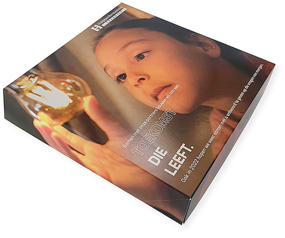 Een schuine bovenaanzicht van een vierkante, full-color bedrukte gebaksdoos met een vast deksel, op maat gemaakt voor Hoppenbrouwers Techniek. De doos, met een afmeting van 270 x 270 x 50 mm, is bedoeld voor een 'Brabantse Broeder' als relatiegeschenk. Het ontwerp toont een artistieke foto van een kind dat met verwondering naar een brandende gloeilamp in hun hand kijkt, wat symbool staat voor innovatie en de toekomst. In de linkerbovenhoek staat het logo van Hoppenbrouwers Techniek. De tekst op de doos luidt: 'Samen met onze partners bouwen wij aan een TOEKOMST DIE LEEFT. Ook in 2022 hopen we weer samen met u antwoord te geven op de vragen van morgen'. Deze luxe verpakking dient als professioneel visitekaartje en versterkt de merkbeleving tijdens het weggeven van gebak.