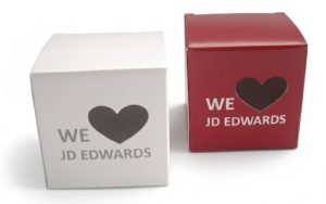 Twee voorbeelden van een bedrukte vensterdoos in de vorm van een kubusdoosje, uitgevoerd in mat wit en dieprood karton. Beide verpakkingen zijn voorzien van een venster in een speciale vorm, namelijk een hartje, dat fungeert als het 'love'-symbool in de tekst "WE [hart-venster] JD EDWARDS". Deze maatwerk kubusdoosjes illustreren hoe een venster in een speciale vorm, zoals een hartje of rondje, extra aandacht trekt en het product direct zichtbaar maakt voor de klant zonder de verpakking te openen. De foto toont de mogelijkheid om logo's en boodschappen te combineren met creatieve venstervormen voor een unieke uitstraling en verbeterde productpresentatie.