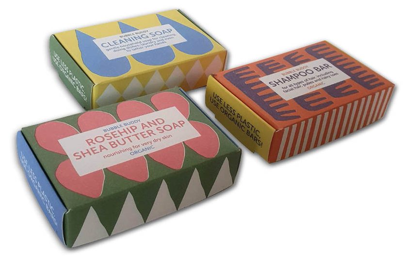 Deze afbeelding toont de veelzijdigheid van de Bubble Buddy-lijn met drie verschillende soap bar verpakkingen. Elk doosje heeft een uniek kleurenschema en patroon (roze bloemvormen, blauwe druppels en oranje golven) die de specifieke functie van de zeep benadrukken. De zijkanten dragen de duidelijke boodschap 'Use less plastic, use organic bars', wat perfect aansluit bij de duurzame uitstraling van het gebruikte hardsized karton. De bedrukking is mat en verzadigd, wat zorgt voor een moderne, kwalitatieve uitstraling in het schap.