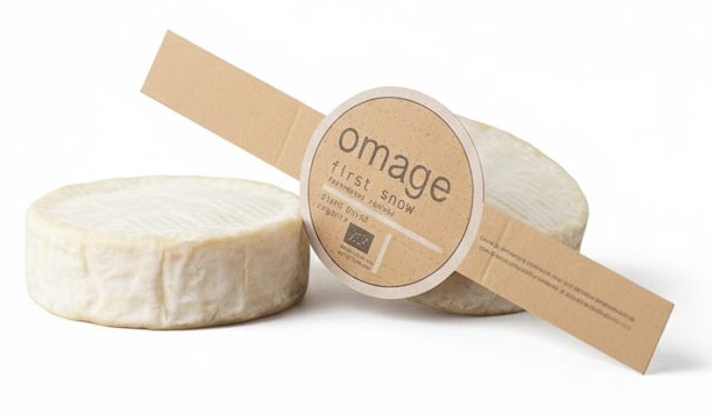 Een ongevouwen, op maat gemaakte kartonnen sleeve voor een veganistisch kaasproduct van het merk 'Omage'. De sleeve is gemaakt van milieuvriendelijk bruin kraft karton met een ronde uitsnede in het midden die fungeert als opvallend presentatievlak voor het logo en productinformatie. De bedrukking bevat termen als 'plant based' en 'organic', samen met het Europese biologische keurmerk. Deze schuifhuls is specifiek ontworpen voor de koeling en is gemaakt van hardsized materiaal dat bestand is tegen temperatuurschommelingen en vocht. Het ontwerp laat zien hoe een sleeve over een deel van het product kan vallen, zodat het product deels zichtbaar blijft voor de klant. De verpakking is bedrukt met plantaardige inkt en is na gebruik volledig recyclebaar via het oud papier.