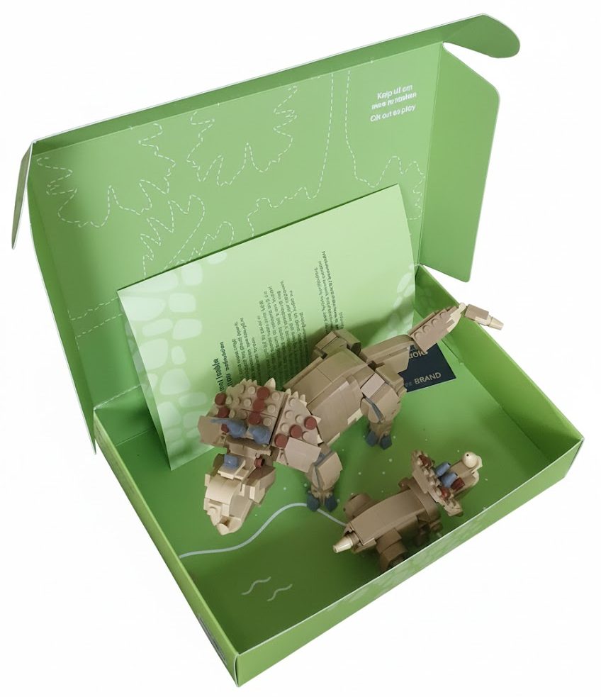 De geopende binnenzijde van een op maat gemaakte groene speelgoeddoos voor een Lego bouwpakket van een Triceratops met jong. De binnenzijde is volledig full-color bedrukt met witte lijntekeningen van bladeren en een stippellijn met de tekst 