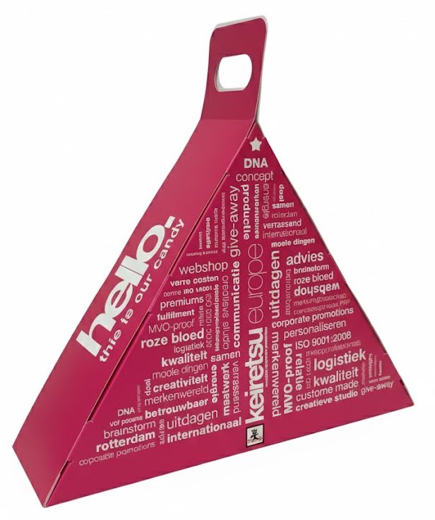 Een opvallende, op maat gemaakte kartonnen doos met handvat in een unieke driehoekige vorm. De verpakking is uitgevoerd in een felle fuchsia roze kleur en volledig bedrukt met een witte woordwolk van relevante zakelijke termen, zoals 'advies', 'creativiteit', 'maatwerk', 'logistiek' en 'personaliseren'. Aan de zijkant staat in grote, verticale witte letters de tekst 'hello! this is our candy'. De doos loopt spits toe naar boven, waar een stevig, geïntegreerd handvat met een ovale uitsparing is verwerkt. Dit ontwerp dient als een originele verpakking voor een give-away of relatiegeschenk en illustreert hoe een doos met handvat op maat gemaakt kan worden om een merk of boodschap krachtig onder de aandacht te brengen bij events en beurzen.