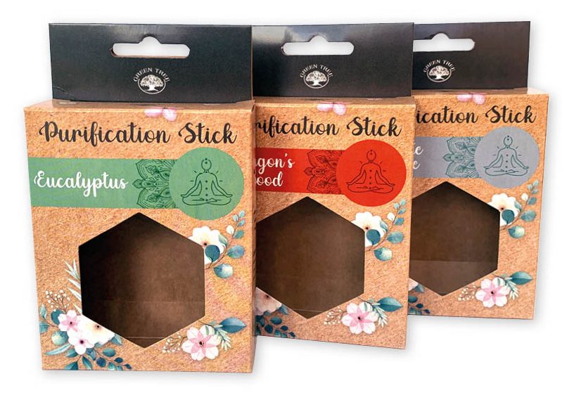 Drie professioneel bedrukte hangende doosjes met venster voor 'Purification Sticks', diagonaal achter elkaar gepresenteerd. De verpakkingen hebben een natuurlijke kurk-look achtergrond versierd met kleurrijke bloemmotieven en een zeshoekig (hexagonaal) uitgestanst venster zonder folie, waardoor het product direct zichtbaar is. Elk doosje heeft een zwart kopstuk met een euroloch sleuf (eurosleuf) voor een geordende presentatie aan een ophanghaak in de winkel. De serie toont verschillende varianten, waaronder 'Eucalyptus' met een groen label en 'Dragon's Blood' met een rood label, elk voorzien van een mediterend icoon. Deze hangverpakking is ontworpen om de klant te verleiden en het merkgevoel te versterken.