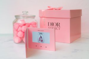 Luxe tweedelige roze geschenkdoos van stevig golfkarton, afgewerkt als dekseldoos met een satijnen lint en zwarte bedrukking met het logo van Dior. Deze duurzame cadeauverpakking wordt gepresenteerd als onderdeel van een giftset met een glazen pot en videokaart, ideaal voor cosmetica en luxe relatiegeschenken.