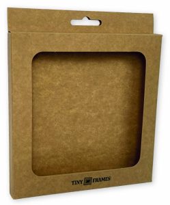 Bedrukte doos van bruin kraft karton met een groot vierkant venster en een euro-slot hangtab (ophangoog) aan de bovenzijde. Het doosje heeft een duurzame, natuurlijke uitstraling en is voorzien van een zwart 'TINY FRAMES' logo, geproduceerd door Drukkerij Van der Louw voor retailproducten in Nederland en België.