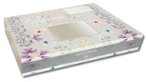 Schuin bovenaanzicht van een luxe, op maat gemaakte kartonnen sleeve met een rechthoekig uitgestanst venster in het midden. De wikkel is voorzien van een verfijnd pastelkleurig bloemendessin met zachte tinten paars, groen en blauw op een lichtgrijze achtergrond. Het ontwerp bevat een subtiel logo ('Stylerone') en een grijze zijbalk met de websitevermelding van de klant. Het venster is nauwkeurig gepositioneerd om het onderliggende product zichtbaar te maken, wat deze schuifhuls ideaal maakt voor luxe textiel, stationery of cadeauverpakkingen. Deze presentatieverpakking combineert hoogwaardige full-color bedrukking met functioneel stanswerk, geproduceerd voor de zakelijke markt in Nederland en België door bedrukte-doosjes.nl.