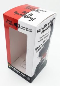 Een rechtopstaande, op maat gemaakte kartonnen speelgoedverpakking met een groot uitgestanst venster aan de voorzijde, gepresenteerd tegen een strakke witte achtergrond. De doos is bedrukt in een heldere rood-witte kleurstelling met speelse zwarte teksten en grafische illustraties. De zijkant toont een silhouet van een knuffel en verwijzingen naar geluidsfuncties, wat de interactieve eigenschappen van de inhoud benadrukt. Het ontwerp heeft een professionele afwerking met scherpe vouwlijnen en een stevige constructie, ideaal voor de presentatie van kinderproducten of promotionele artikelen in de retail. De verpakking combineert visuele aantrekkingskracht met functionele productbescherming.