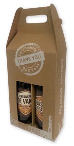 Luxe, gepersonaliseerde 2-pack bierverpakking van stevig, natuurlijk bruin vouwkarton voor twee flesjes (33cl). Het doosje heeft een robuuste 'kraft' look met een ingestanste handgreep aan de bovenzijde voor veilig draaggemak. Op de voorzijde staat een witte, ruitvormige stamp met de tekst 'THANK YOU' en 'BAS VANS'. Aan de zijkant is de tekst 'BAS VANS' zichtbaar in donkerblauwe, gestileerde letters. De onderrand heeft een gedetailleerde witte silhouette van een stadsgezicht met grachten, gebouwen, een kerktoren en een windmolen. Het model heeft aan de voorzijde een groot, uitgestanst venster waardoor de bieretiketten van beide flesjes direct zichtbaar zijn. Het linker flesje toont het donkerblauwe etiket van 'BROUWERIJ DE VAN / AMBACHTELIJK BIER SINCE 1966', en het rechter flesje het etiket van 'BAS BEST / 60 JAAR VAKMANSCHAP'. Een perfect voorbeeld van een gepersonaliseerde 33cl flesverpakking met hoogwaardige bedrukking direct op het karton.