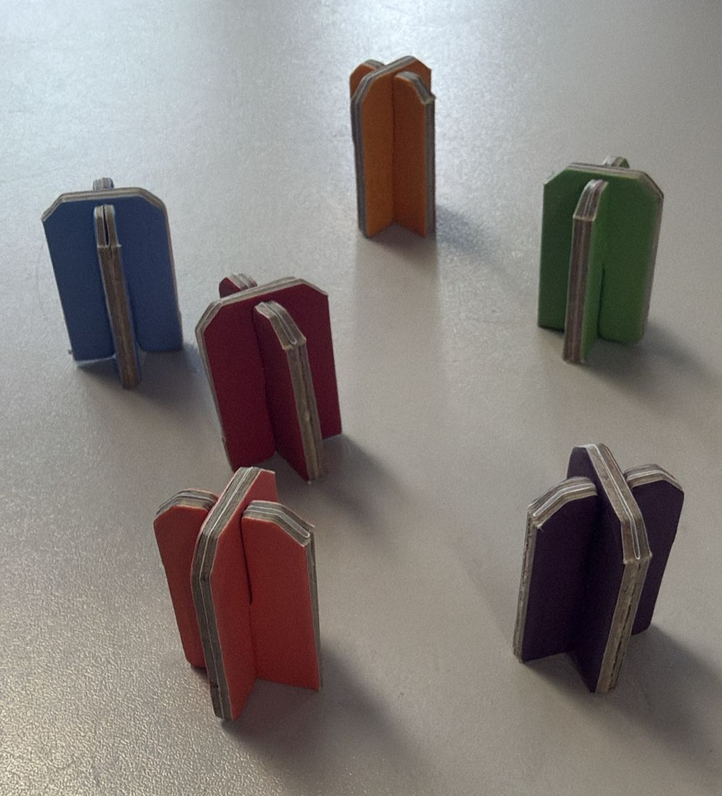 Zes kruislingse spelpionnen van dik grijskarton met een full-color bedrukking in blauw, oranje, groen, rood, koraal en paars. De op maat gemaakte kartonnen speelfiguren (25x25x35 mm) staan als spelaccessoires opgesteld op een egale ondergrond voor een op maat geproduceerde speldoos of bordspel.