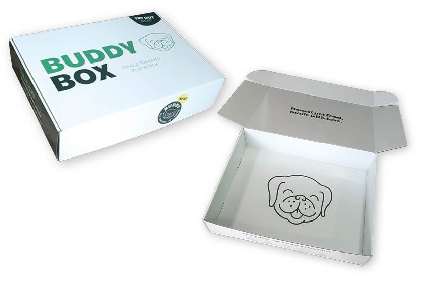 Twee witte, bedrukte verzenddozen van stevig golfkarton met een klepsluiting, voor natuurlijke hondenvoeding. Links op de foto staat een gesloten doos met op de deksel in grote, opvallende donkergroene en zwarte letters de naam met daaronder in een dun, elegant groen lettertype de tekst
