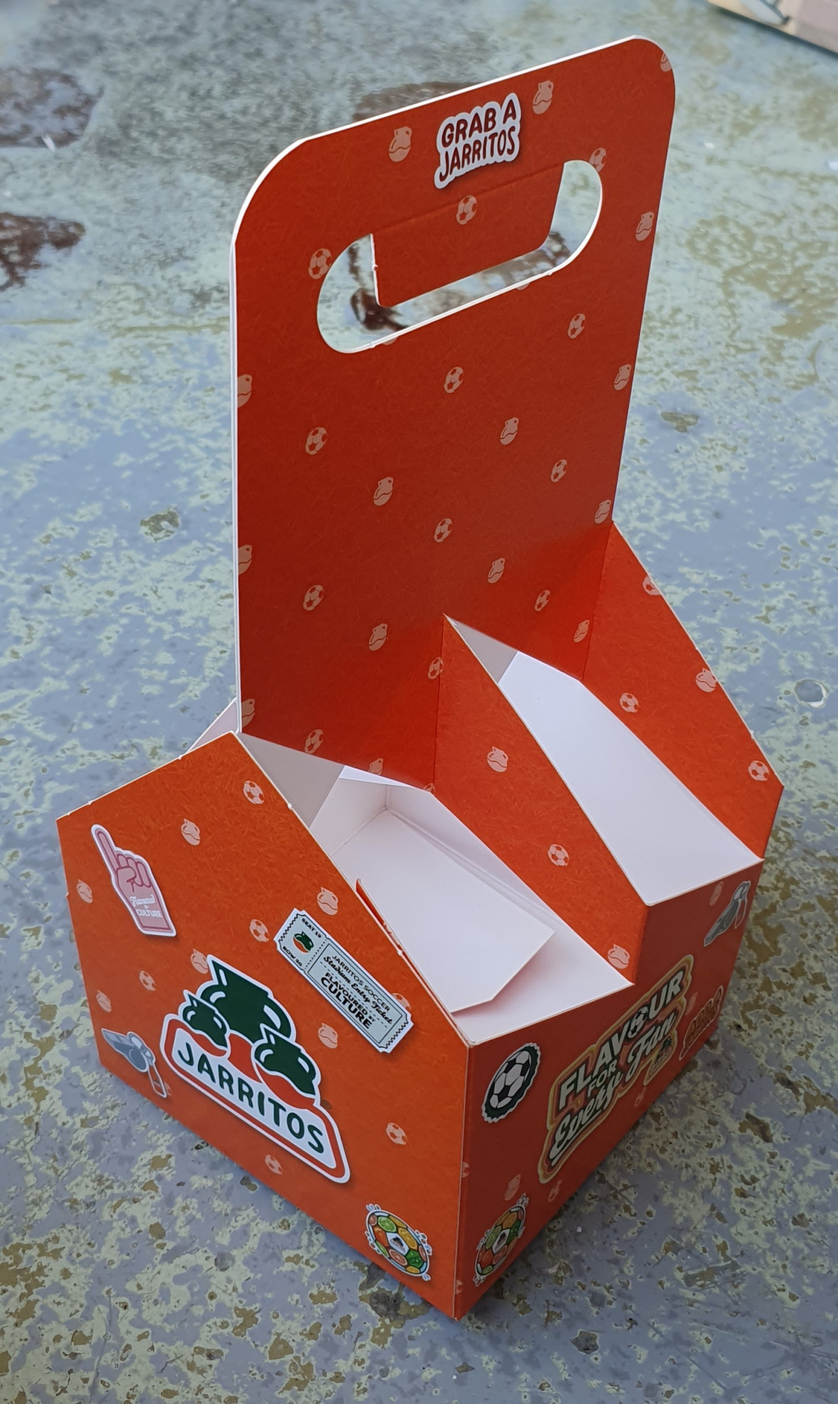 Bedrukte 4-pack flesverpakking van stevig vouwkarton voor vier flesjes van 33cl, uitgevoerd in opvallend oranje met full color bedrukking. De houder heeft een hoog uitgestanst handvat en een open vakverdeling voor vier 33cl flesjes. De voorzijde toont het Jarritos-logo met voetbal- en cultuurstickers en de tekst 'Grab a Jarritos' bovenaan. Een levendig voorbeeld van een gepersonaliseerde 33cl 4-pack flesverpakking met hoogwaardige full color bedrukking direct op het vouwkarton.