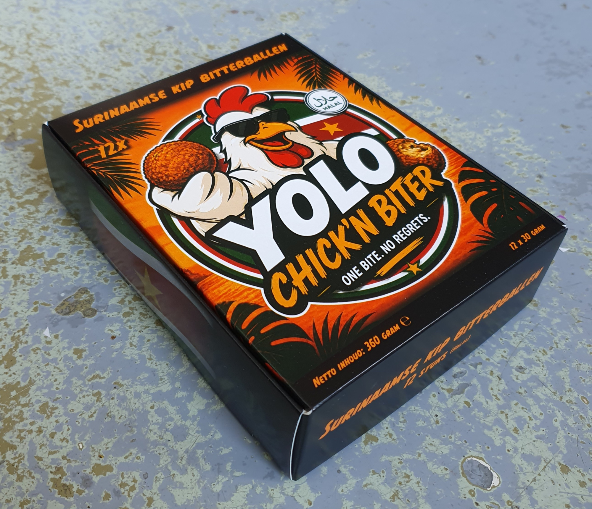 Een felgekleurde bedrukte doos voor diepvries bitterballen van het merk YOLO Chick'n Biter, gemaakt voor NSS-Food uit Purmerend. De zwarte doos met oranje tropische bedrukking toont een illustratie van een haan met zonnebril en bitterbal, met de tekst 'Chick'n Biter – One bite. No regrets.' en een halal-keurmerk. De verpakking bevat 12 Surinaamse kip bitterballen van 30 gram (360 gram totaal), geschikt voor de airfryer. De doos is gemaakt van koeling-geschikt karton dat de bitterballen optimaal beschermt in de diepvries, terwijl het speelse ontwerp zorgt voor herkenning in het schap.