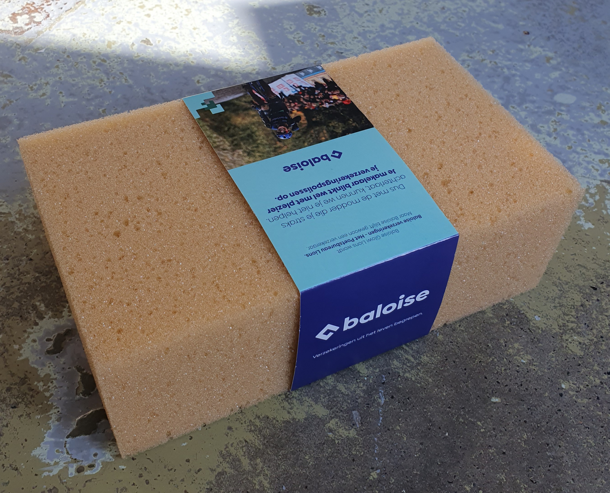 Bedrukte kartonnen wikkel in full color om een grote sponsblok, uitgevoerd voor Baloise verzekeringen België. De wikkel is gedrukt in het kenmerkende blauw en lichtgroen van Baloise, met het logo en de tagline 'Verzekeringen uit het leven begrepen' op de voorzijde. De achterzijde toont een foto van een wielrenwedstrijd met de tekst 'Dus met de modder die je straks achterlaat, kunnen we je niet helpen. Maar je makelaar blinkt wel met plezier je verzekeringspolissen op.' — een verwijzing naar de naamsverandering van Baloise Glowi Lions naar Het Poetsbureau Lions. Een creatief voorbeeld van een bedrukte wikkel als relatiegeschenk bij een rebranding, geproduceerd door Drukkerij Van der Louw in Berkel en Rodenrijs.