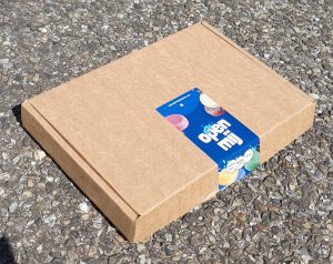 Een gesloten, milieuvriendelijke bruine brievenbusdoos, gepresenteerd op een grove grindondergrond. De doos is beveiligd met een levendige, blauwe sluitsticker met de witte, vrolijke tekst 'open mij' en het logo. De sticker toont ook illustraties van diverse kleurrijke shampoo bars en een kleiner wit label met de claims: 'Gemaakt in NL', '100% plasticvrij' en 'Vegan'. De foto is genomen vanuit een verhoogd hoekperspectief, met duidelijk gedetailleerde karton- en grindtexturen onder helder daglicht.