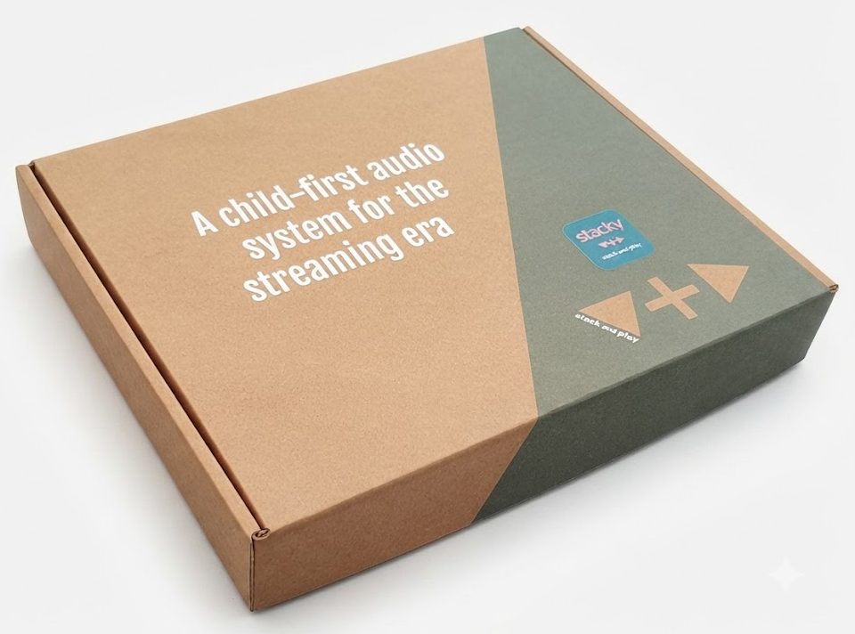 Professionele productfoto van een bedrukte verzenddoos van bruin golfkarton voor een kinderspeelgoed of audiosysteem genaamd 'stacky'. De doos heeft een strakke, eigentijdse opdruk met witte typografie aan de linkerzijde ('A child-first audio system for the streaming era') en een contrasterend groen vlak met een speels logo en bedieningssymbolen aan de rechterzijde. De verpakking is vrijstaand gepresenteerd tegen een egale, lichte achtergrond om de milieuvriendelijke kartonstructuur en hoogwaardige bedrukking te benadrukken.