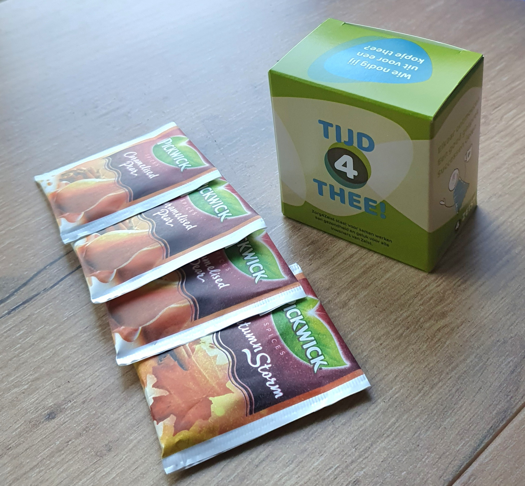 Een groen gepersonaliseerd theedoosje met de tekst 'TIJD 4 THEE!' en een uitnodigende vraag op de bovenkant, stijlvol gepresenteerd op een houten ondergrond naast vier geportioneerde Pickwick theezakjes in herfstige smaken als Caramelised Pear en Autumn Storm. In het doosje gaan 6 zakjes thee.