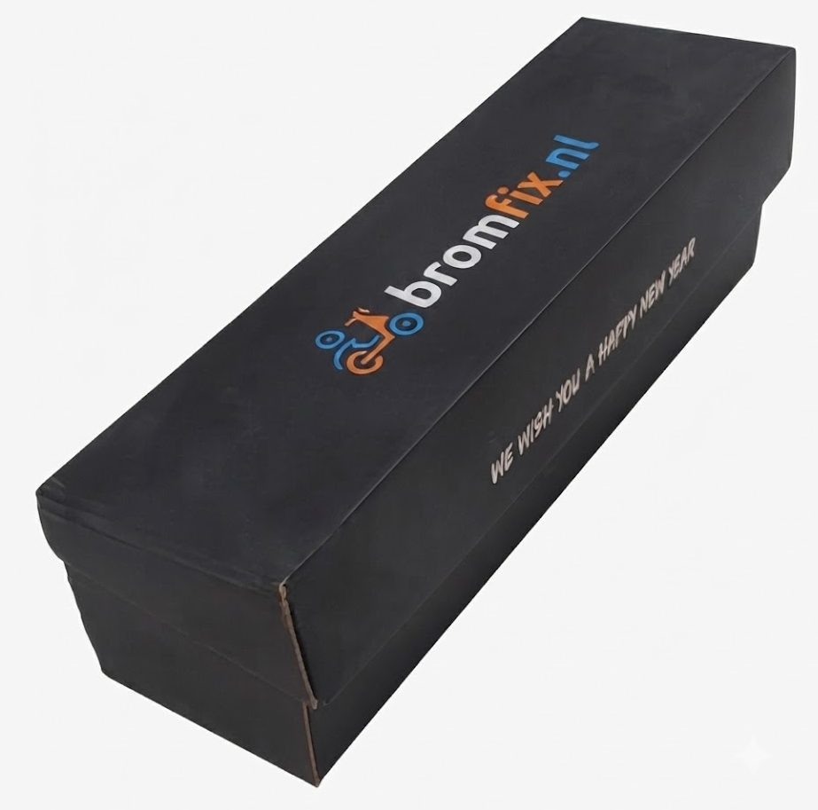 foto van een premium zwarte geschenkdoos voor een enkele wijnfles, De foto toont luxe doos volledig gesloten, eveneens in een hoge hoek. Op het deksel bevindt zich het meerkleurige logo van Bromfix.nl, met blauwe en oranje typografie vergezeld van een kleine, gestileerde oranje scooter-icoon. De verpakking straalt kwaliteit uit voor een gedenkwaardig relatiegeschenk.